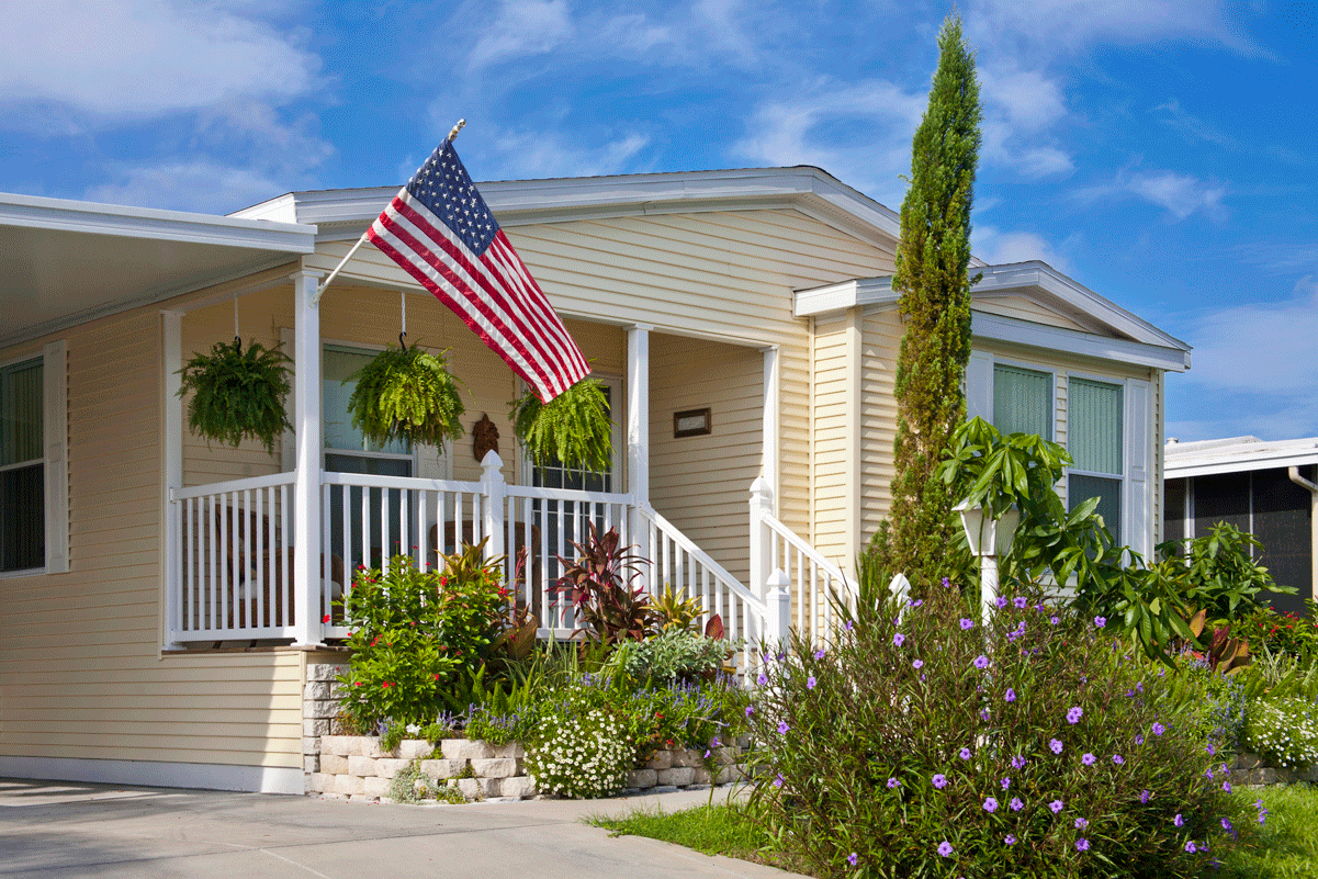 Front entrance image of one-level manufactured home with garden and United States flag.